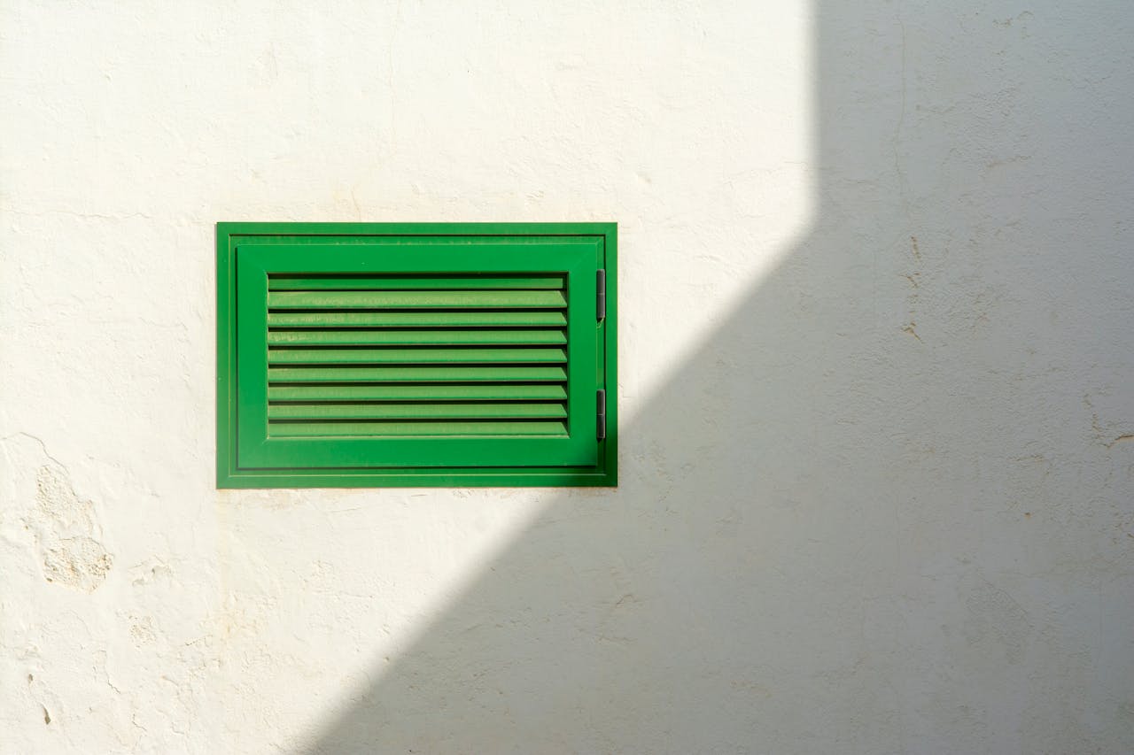 Green vent on a white wall with sharp sunlight casting a shadow, creating a minimalist urban scene.